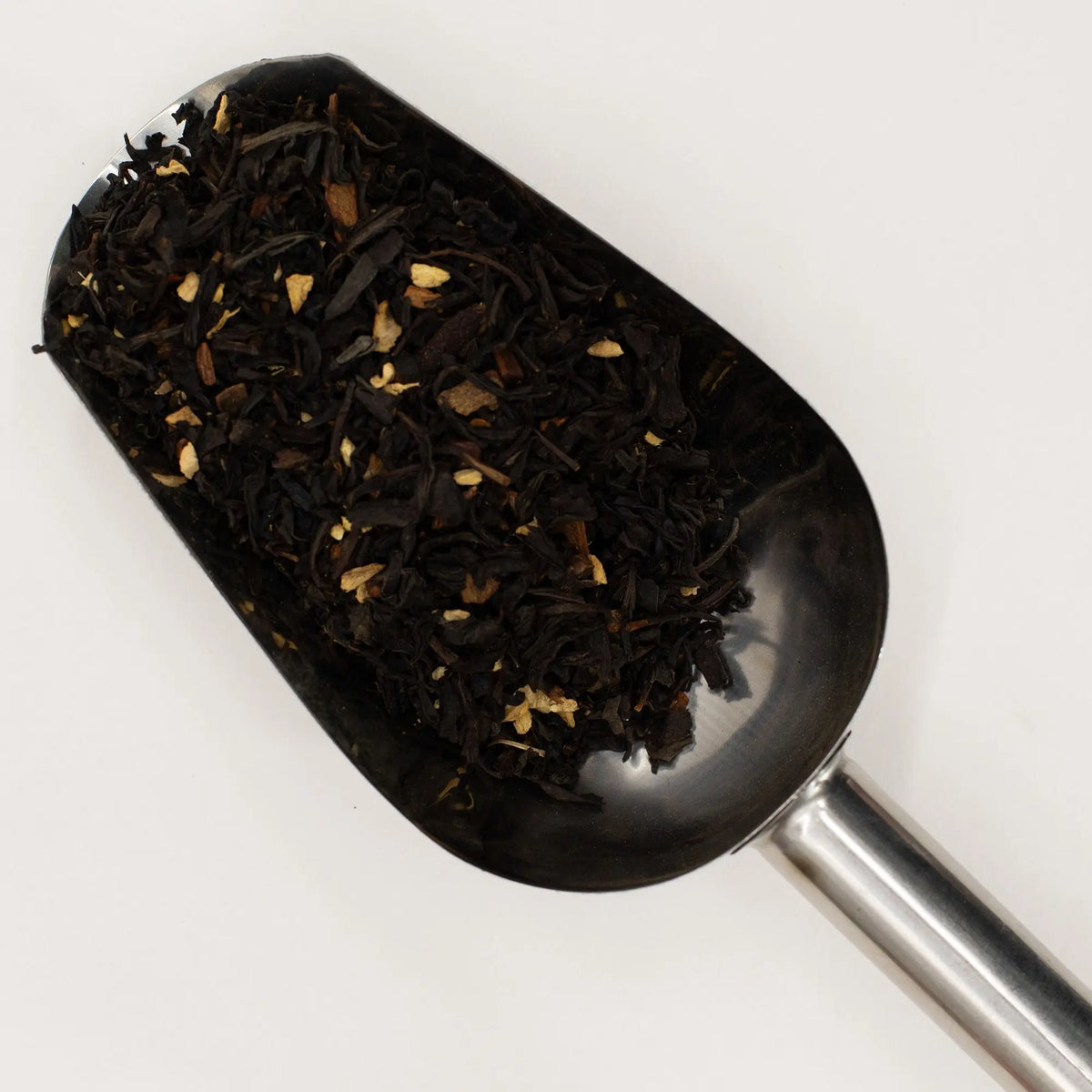 Pala de metal con té negro orgánico y trozos de especias sobre fondo blanco neutro.