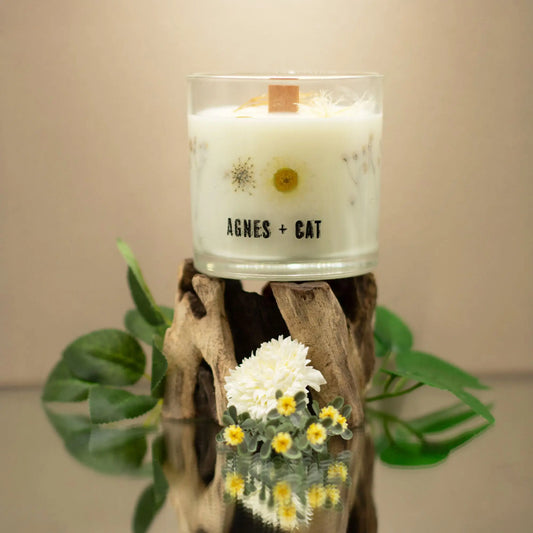Vela blanca decorativa con flores secas, envase de vidrio, marca Agnes + Cat, sobre base de madera natural.