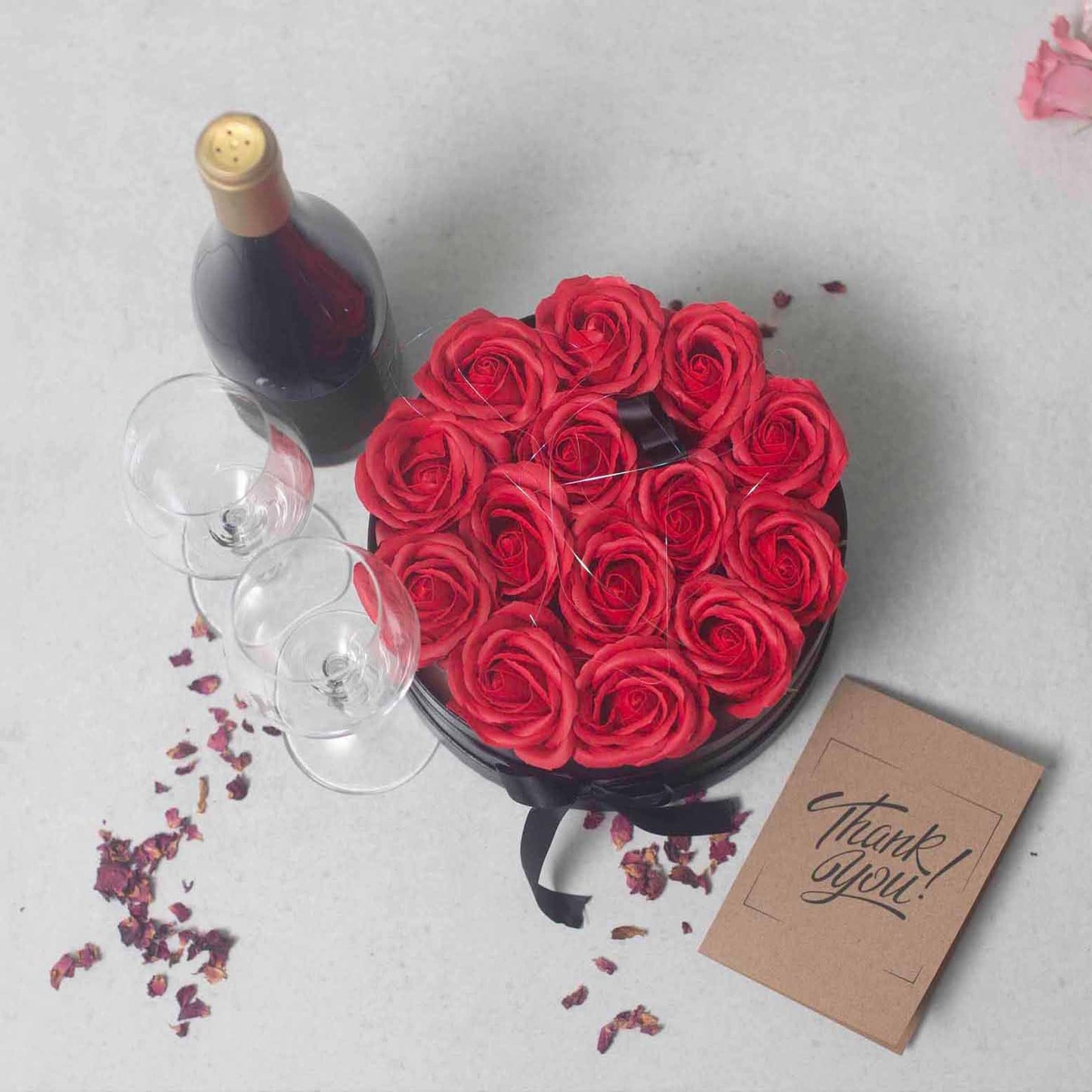 Caja redonda negra con 14 rosas rojas de jabón, junto a una botella de vino, dos copas y una tarjeta de agradecimiento.