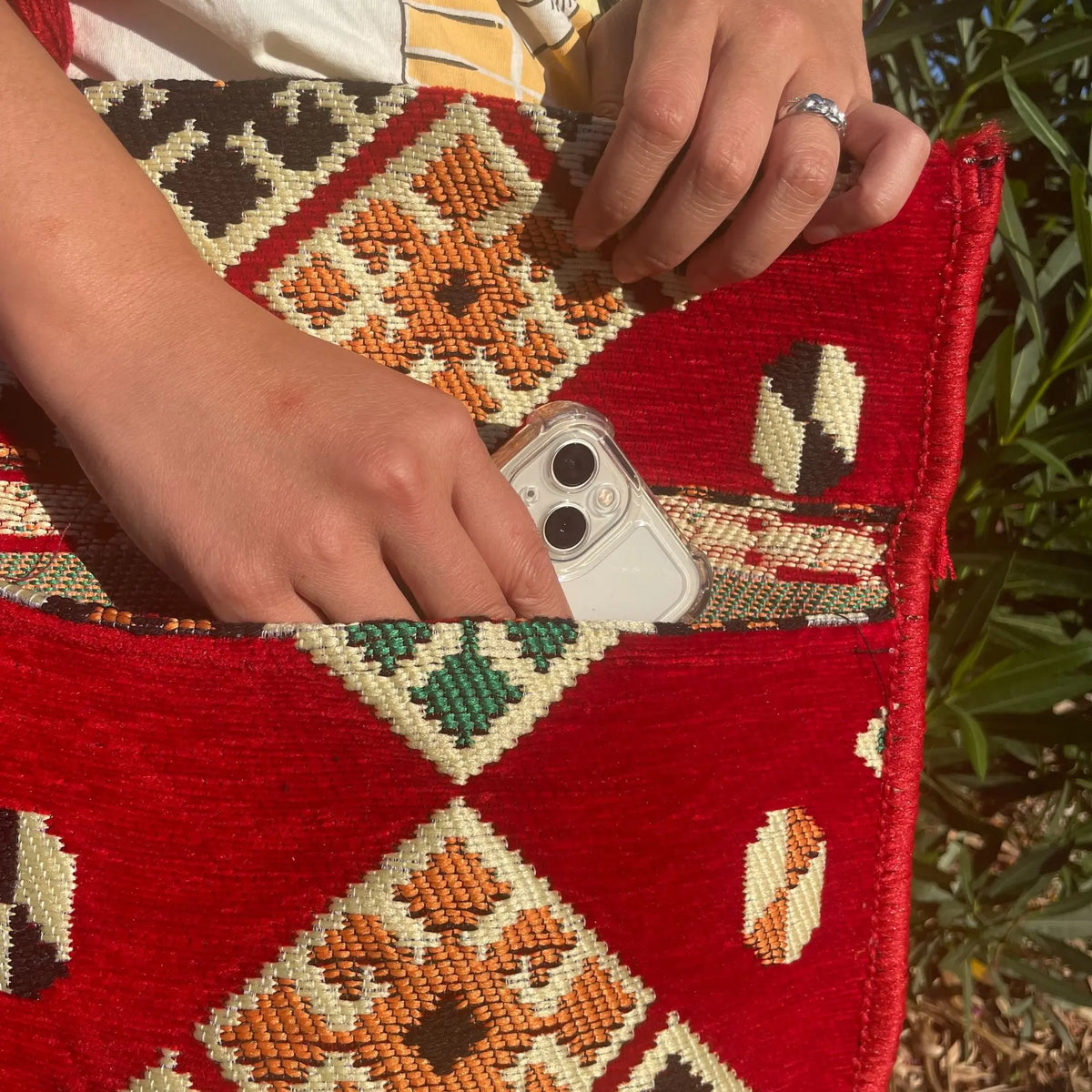 Bolso bandolera de tela roja con motivos geométricos bordados, manos introduciendo un móvil en un bolsillo exterior.