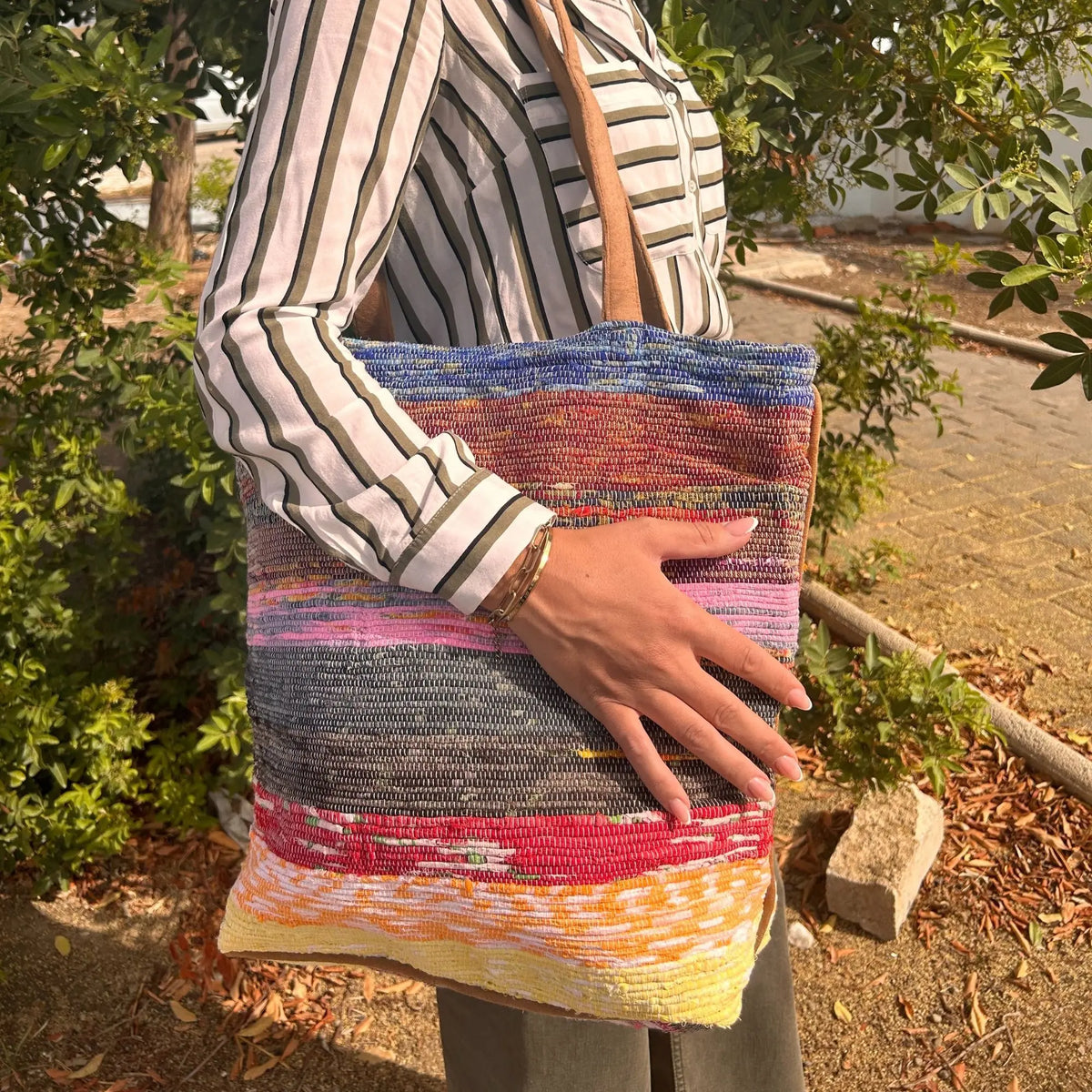 Bolso grande de tela tejida reciclada en franjas multicolores, con asas de cuero marrón, al hombro.