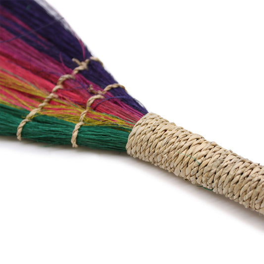 Escobilla artesanal de colores vibrantes: púrpura, verde y rosa, con mango de mimbre trenzado.