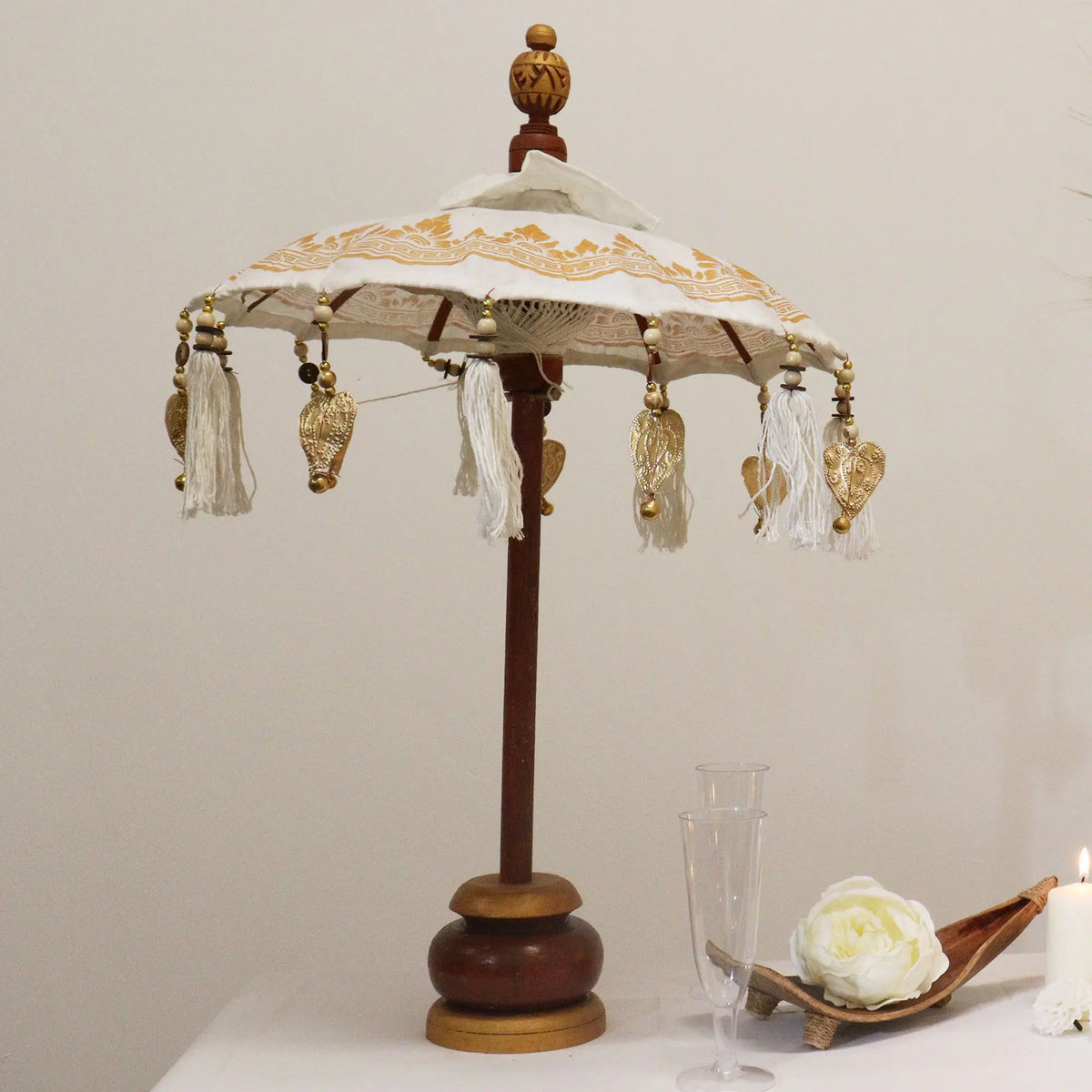 Sombrilla decorativa de Bali, lona blanca con detalles dorados, colgantes y base de madera sobre fondo blanco.