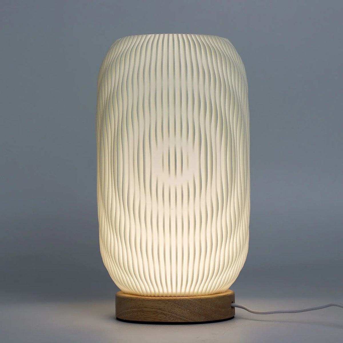 Lámpara de mesa moderna con pantalla cilíndrica de estrías verticales blancas y base de madera circular.