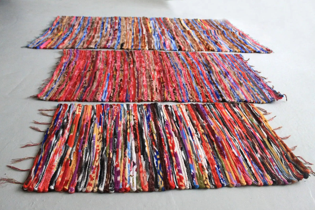 Tres alfombras rectangulares de trapo artesanal indio, tejidas con tiras de tela multicolor y flecos en los extremos.