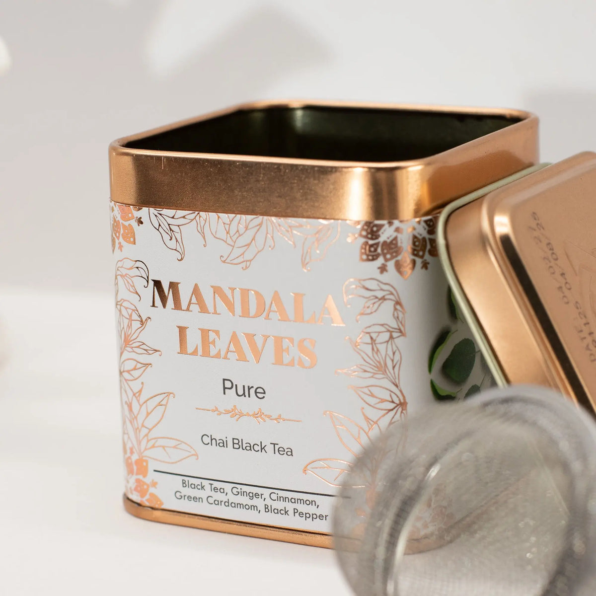 Lata cuadrada de metal color blanco y cobre con diseño floral, junto a un infusor de té sobre fondo blanco.