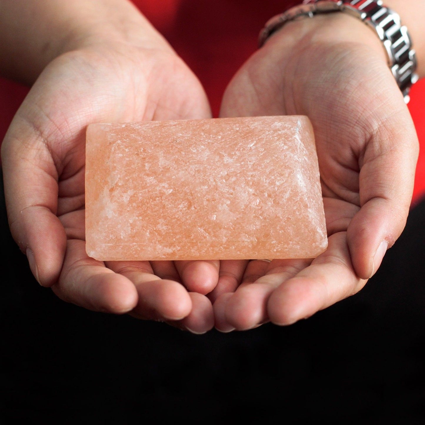 Manos sosteniendo una barra rectangular de sal mineral del Himalaya color rosado con textura cristalina y rugosa.