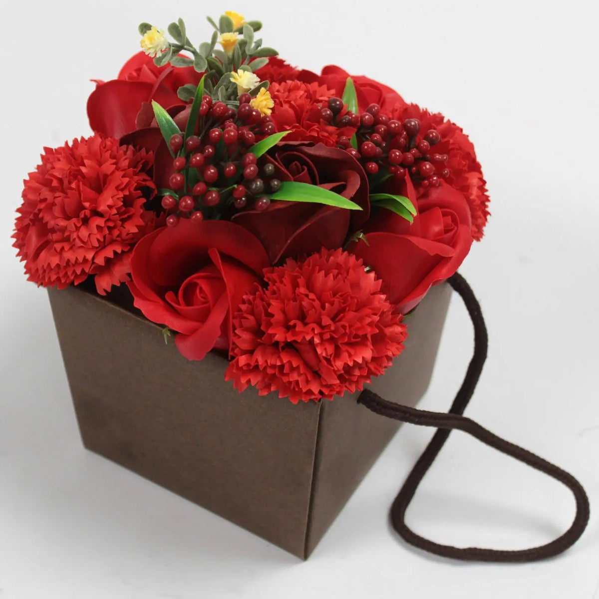 Bouquet de rosas y claveles rojos de jabón en una caja de regalo marrón con asa, decorado con pequeñas bayas y flores.