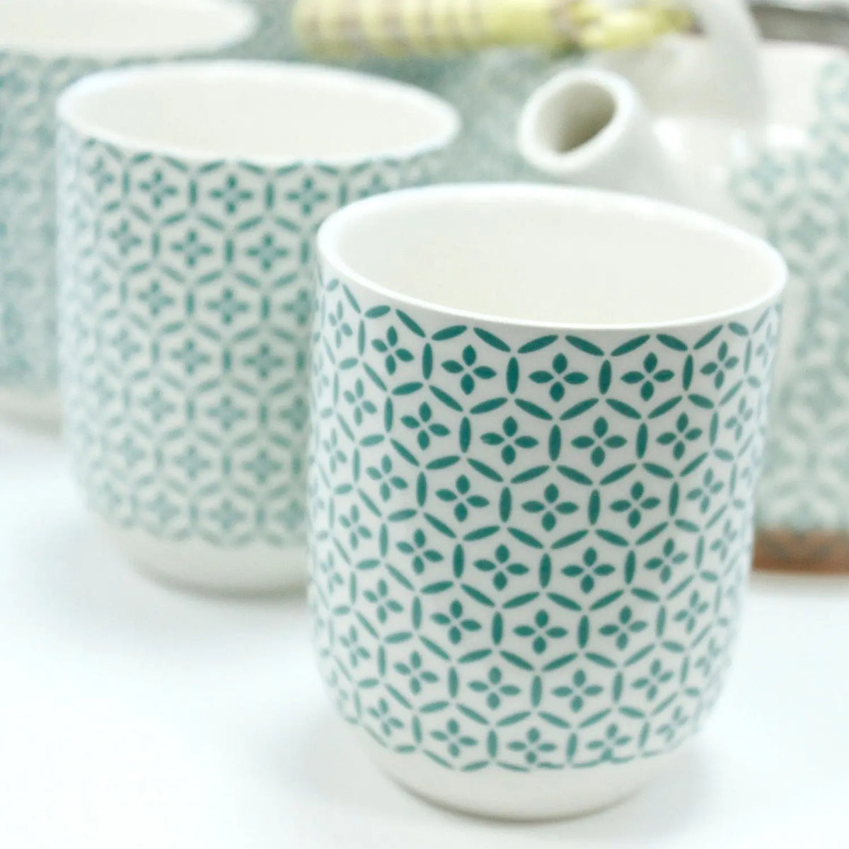 Juego de cerámica blanca con tazas cilíndricas y tetera decoradas con un patrón de mosaico floral en color verde.