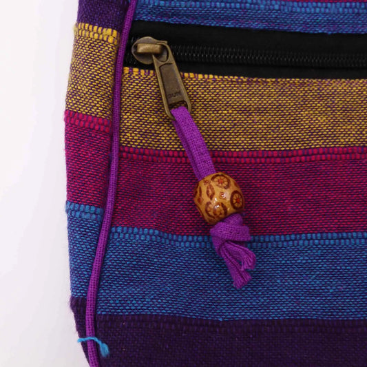Bolso artesanal de tela tejida a rayas coloridas con cremallera, tirador de hilo morado y cuenta decorativa de madera.
