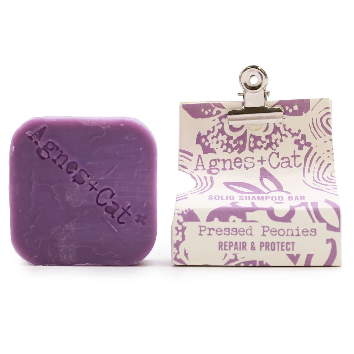 Champú sólido morado Agnes + Cat de peonías, cuadrado, junto a su envoltorio de papel estampado con pinza metálica.