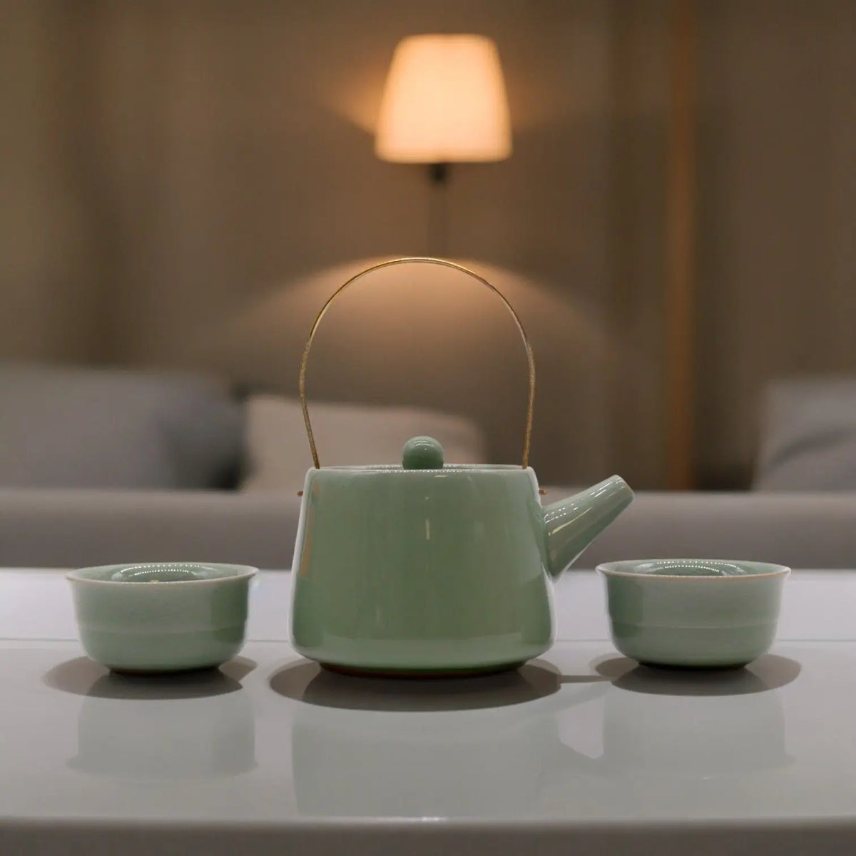Set de té Ancient Wisdom: tetera cerámica verde con asa metálica y dos tazas a juego sobre mesa blanca.