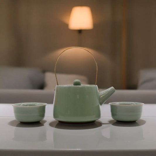 Set de té Ancient Wisdom: tetera cerámica verde con asa metálica y dos tazas a juego sobre mesa blanca.