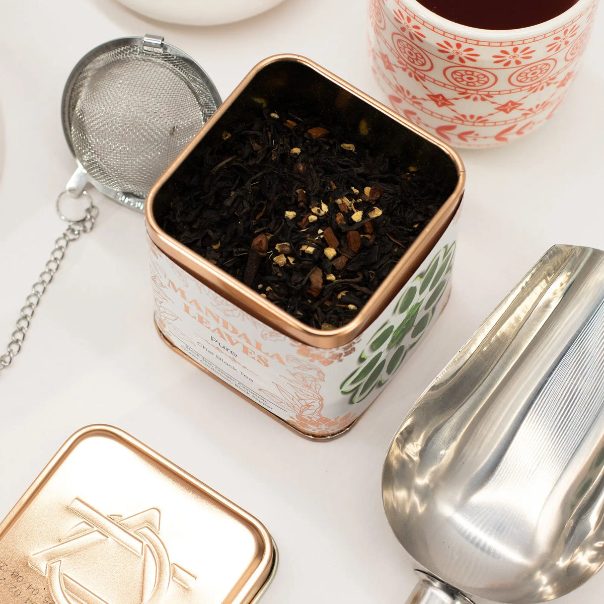 Lata cuadrada de té negro chai con especias, junto a una pala de metal, un infusor y una taza decorada sobre fondo blanco