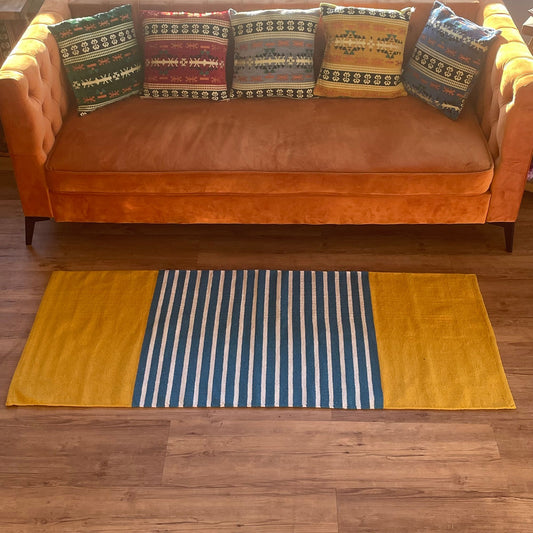 Alfombra de algodón rectangular con franjas verticales azules y blancas en el centro y laterales amarillos lisos.