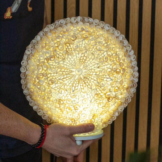Lámpara boho redonda de 30 cm hecha con conchas marinas blancas dispuestas en un patrón floral iluminado.