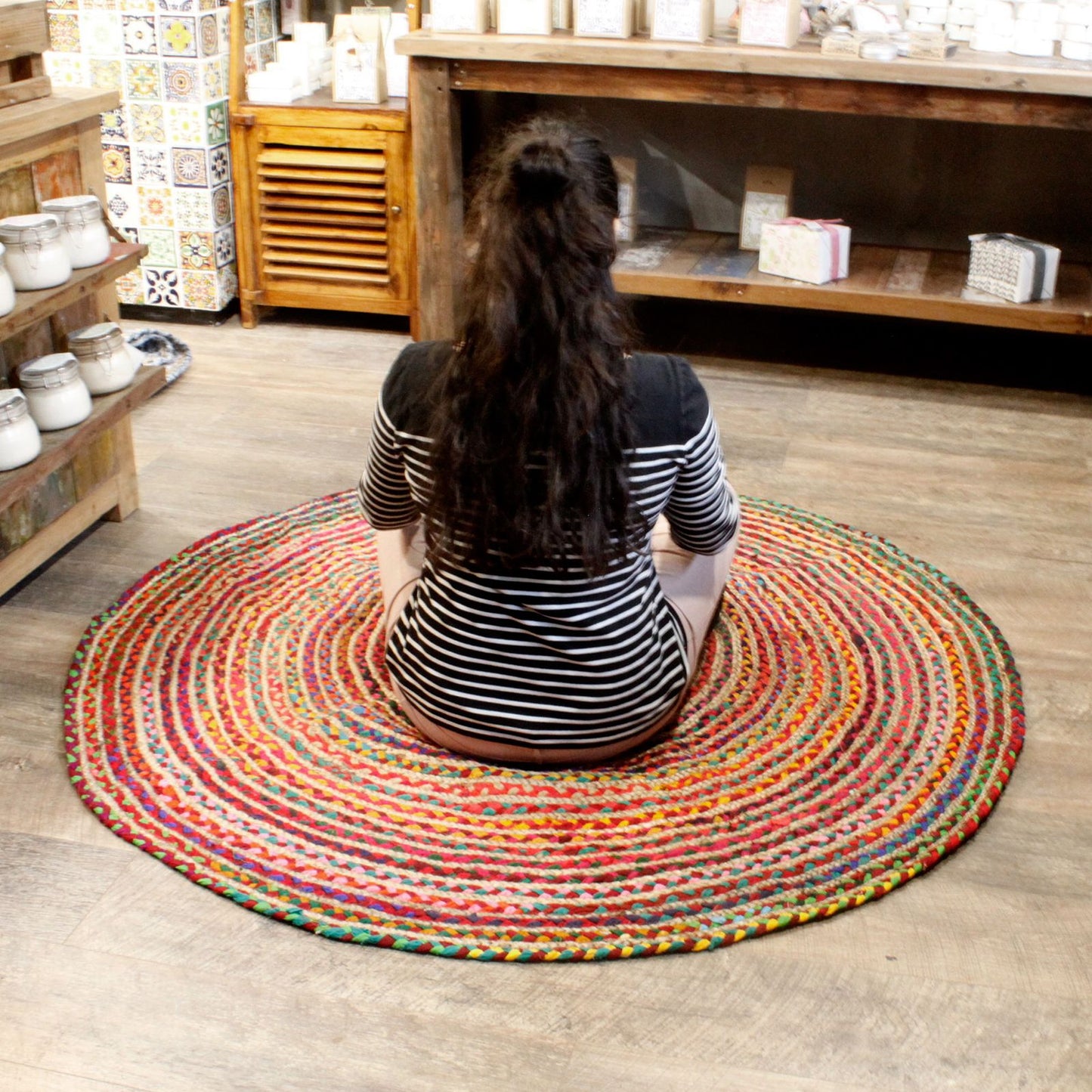 Alfombra redonda de yute y algodón reciclado multicolor, tejida en espiral, sobre suelo de madera en una tienda.