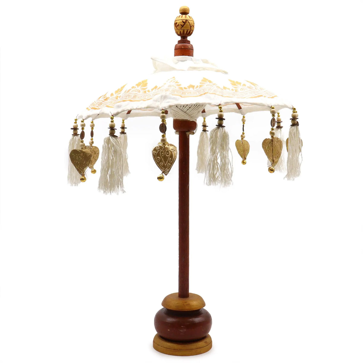 Sombrilla decorativa balinesa de lona blanca con detalles dorados, borlas y adornos metálicos sobre base de madera.