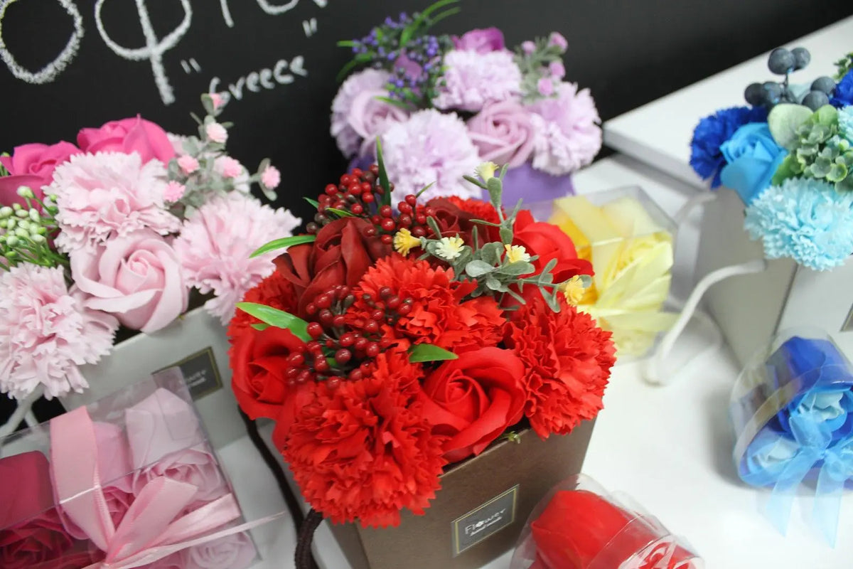 Bouquet de flores de jabón en tonos rojos, rosas y azules, presentadas en cajas decorativas con follaje artificial.