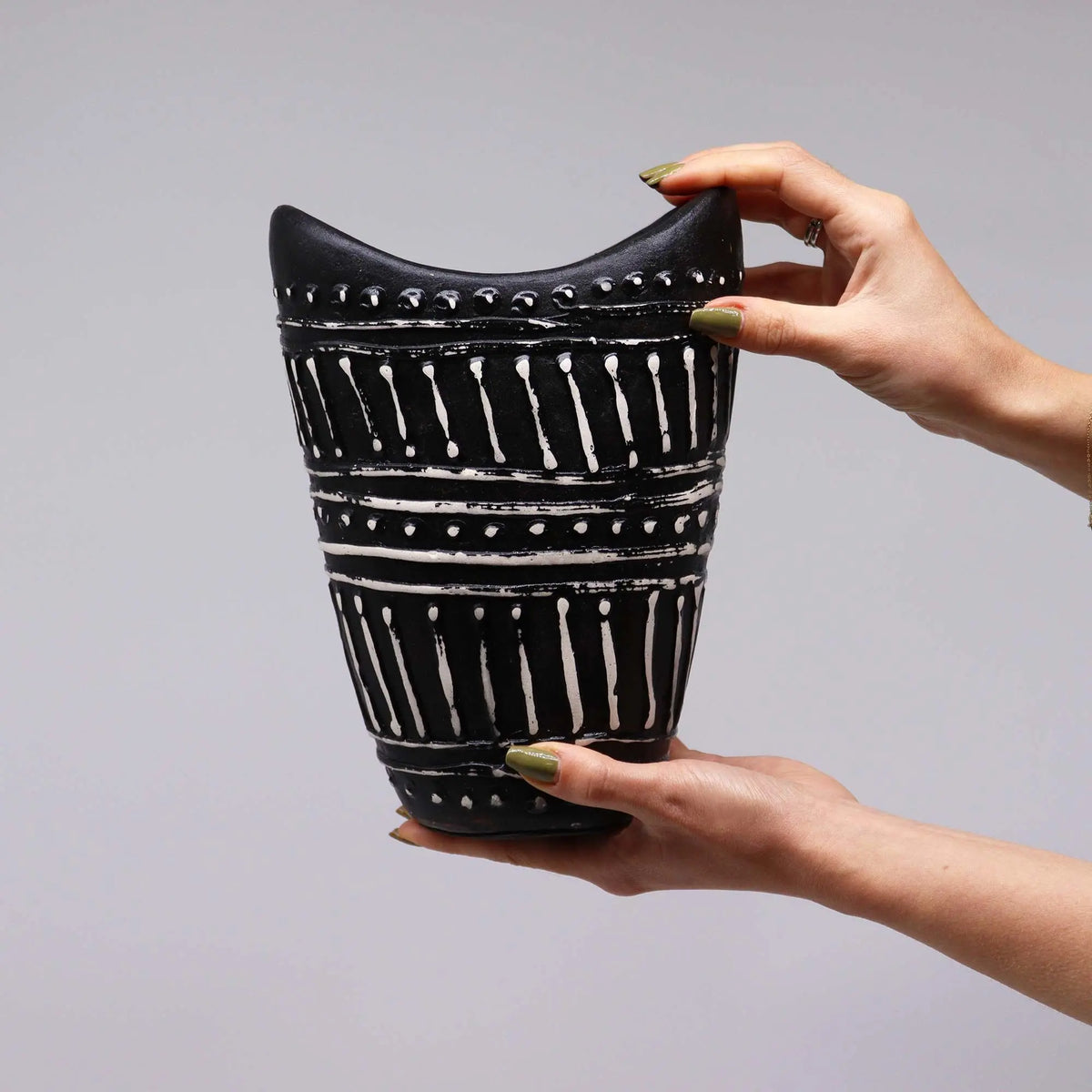 Jarrón de cerámica negro con forma cóncava y patrones geométricos tallados en color blanco, sostenido por dos manos.