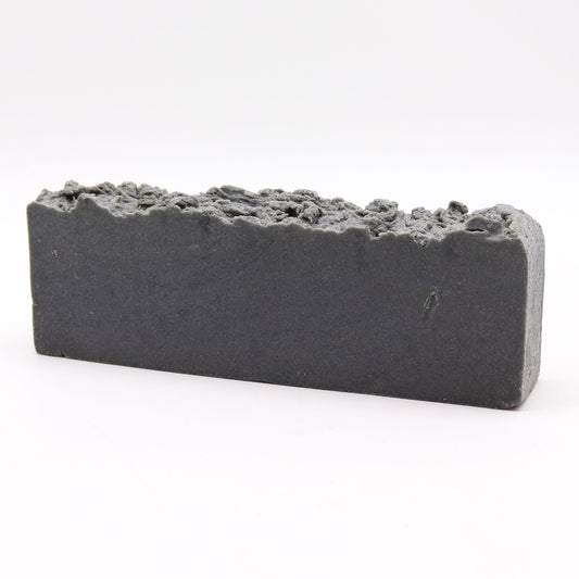 Barra rectangular de jabón artesanal gris oscuro con textura rugosa y granulada en la parte superior. Fondo blanco.