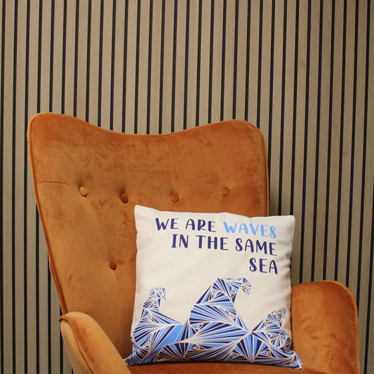 Funda de cojín de algodón color natural con estampado de olas geométricas azules y frase en gris sobre sillón naranja.