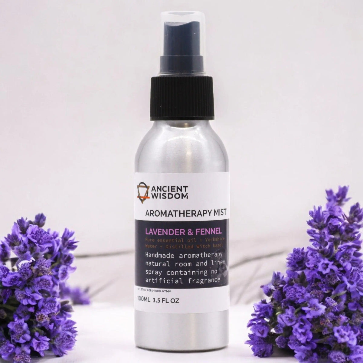 Frasco de aluminio con atomizador negro de bruma de lavanda e hinojo, flanqueado por flores de lavanda sobre fondo claro.
