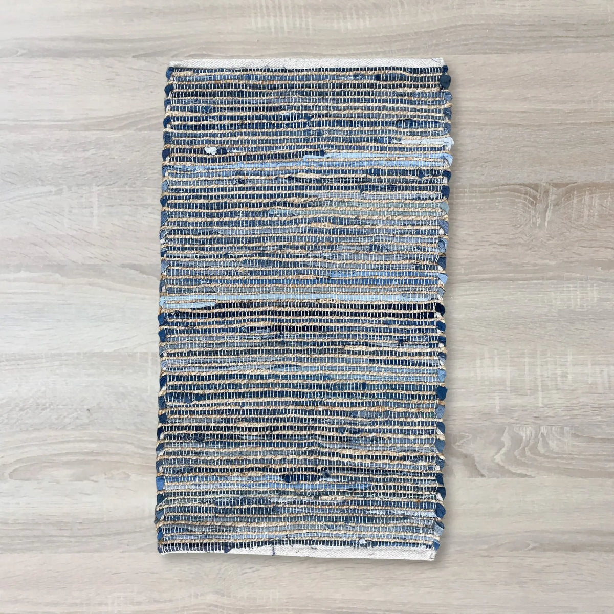 Alfombra rectangular de 50x90 cm tejida en yute y tiras de algodón reciclado en tonos azules sobre fondo de madera.