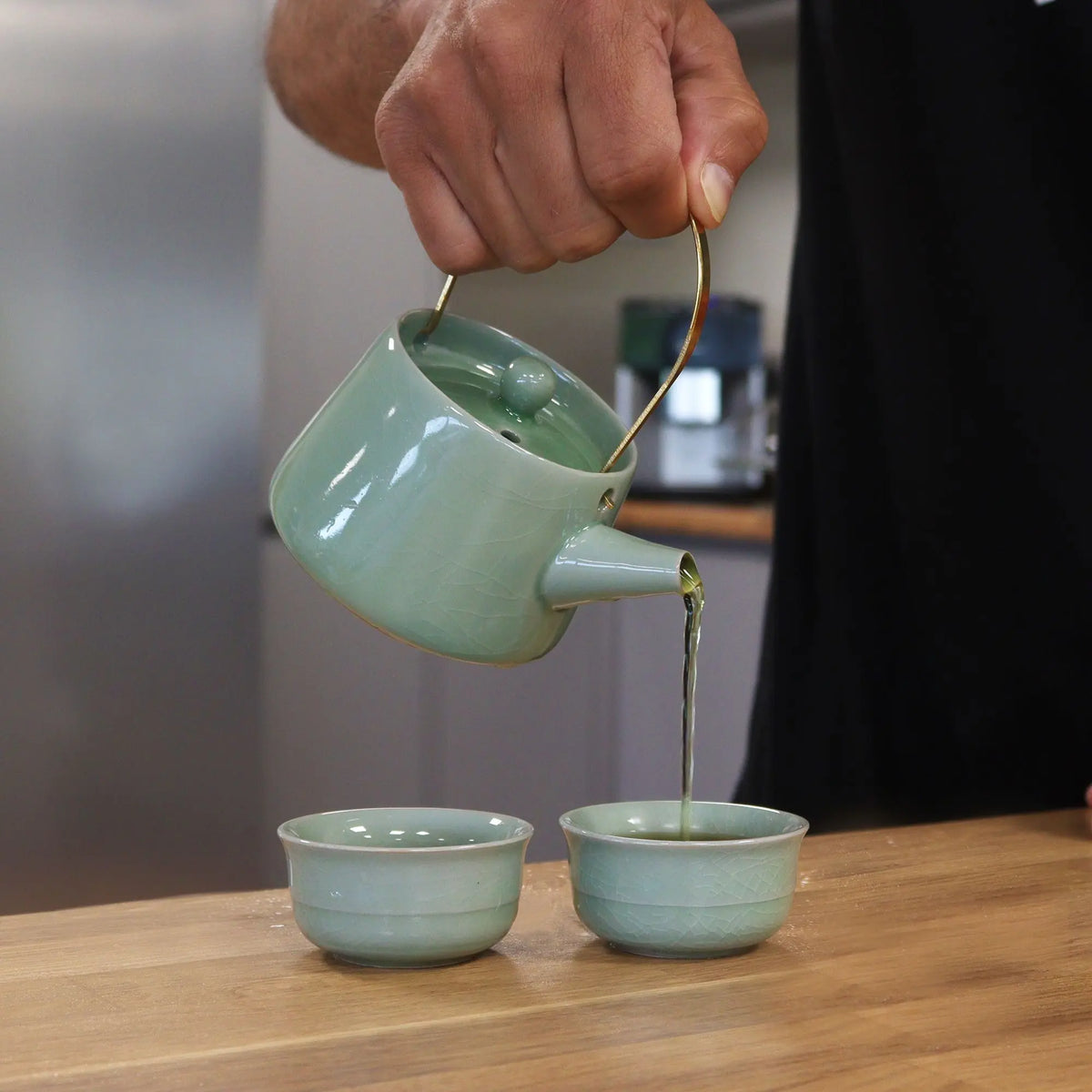 Tetera de cerámica verde con asa dorada sirviendo té en dos tazas pequeñas a juego sobre mesa de madera.