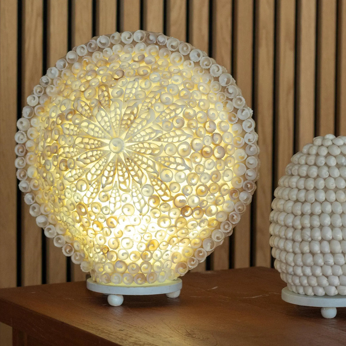 Lámpara redonda boho de 30 cm hecha con pequeñas conchas blancas dispuestas en un patrón floral iluminado desde atrás.