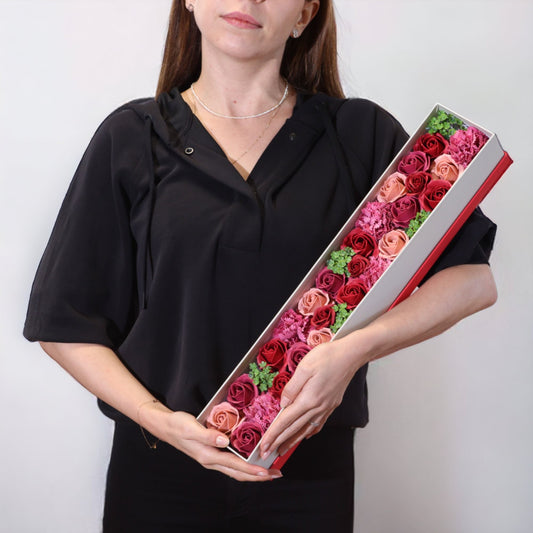 Mujer sosteniendo una caja larga rectangular con rosas de jabón en tonos rojos, rosas y detalles verdes decorativos.