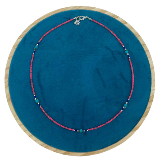 Collar artesanal de cuentas de coral rosa y turquesas con detalles en plata sobre base circular de terciopelo azul.