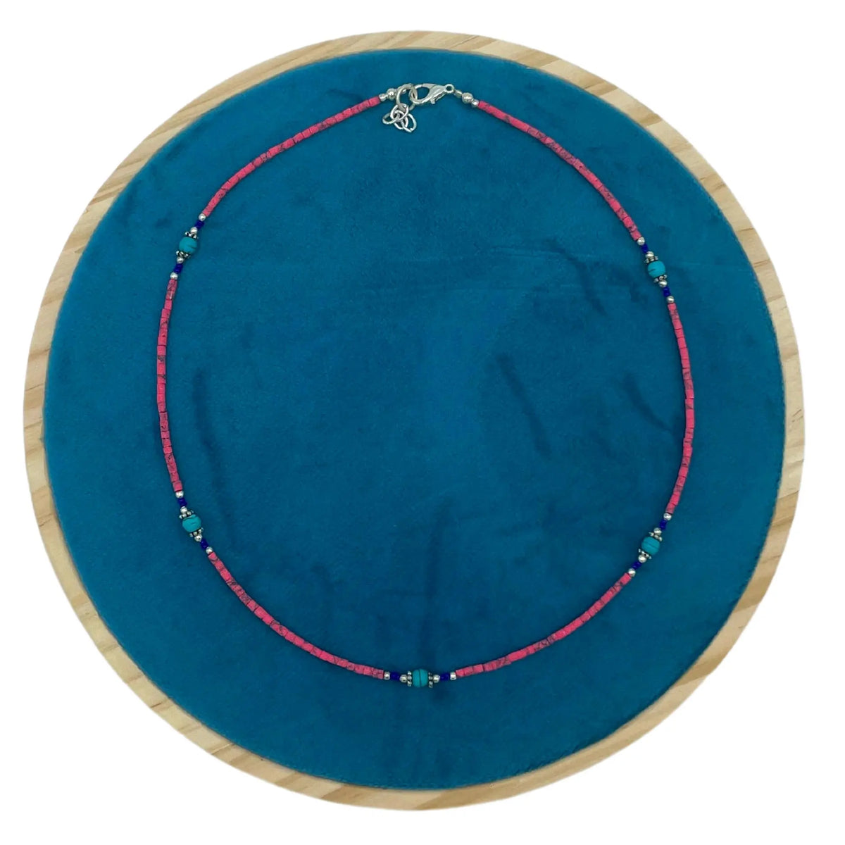Collar artesanal de cuentas de coral rosa y turquesas con detalles en plata sobre base circular de terciopelo azul.