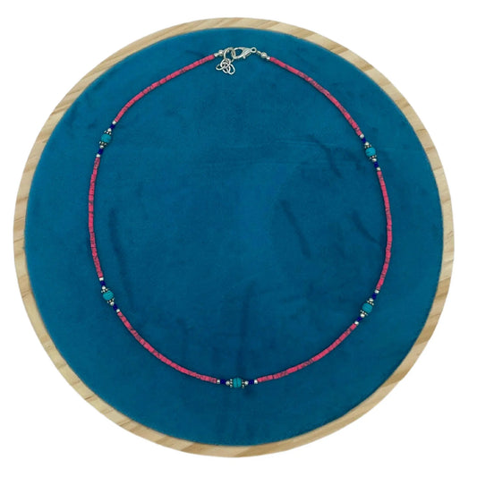 Collar artesanal de cuentas de coral rosa y turquesas con detalles en plata sobre base circular de terciopelo azul.