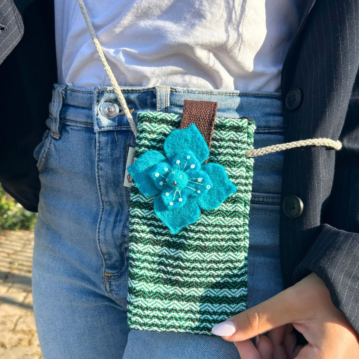 Bolso porta móvil de tela verde a rayas con una flor de fieltro azul brillante, colgado de una cuerda clara.
