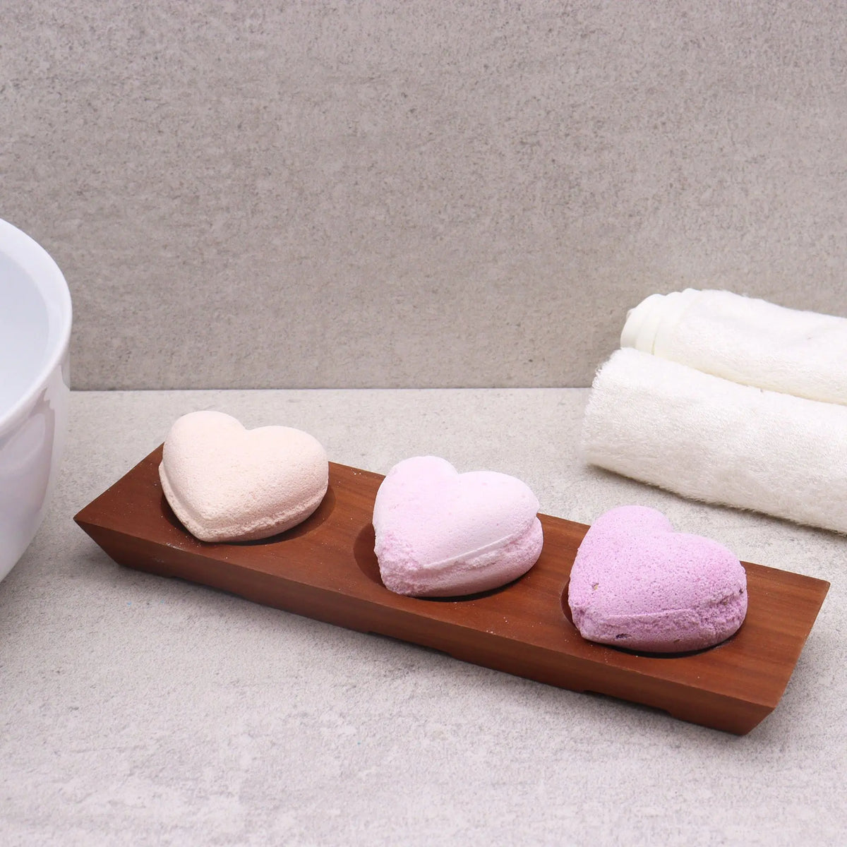 Jabonera rectangular de madera oscura con tres huecos que contienen tres bombas de baño en forma de corazón.