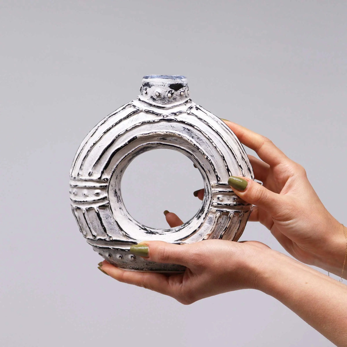 Jarrón de cerámica crema en forma de donut con textura rústica y detalles lineales oscuros, sostenido por dos manos.