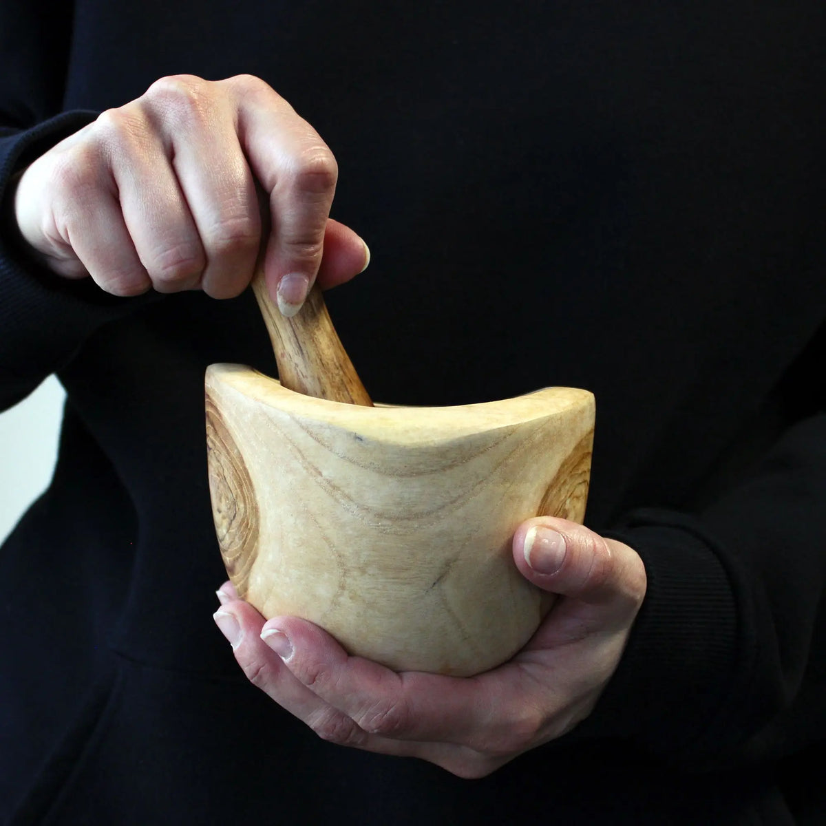 Manos sosteniendo un mortero y maja de madera de teca clara con veteado natural, estilo artesanal y orgánico.