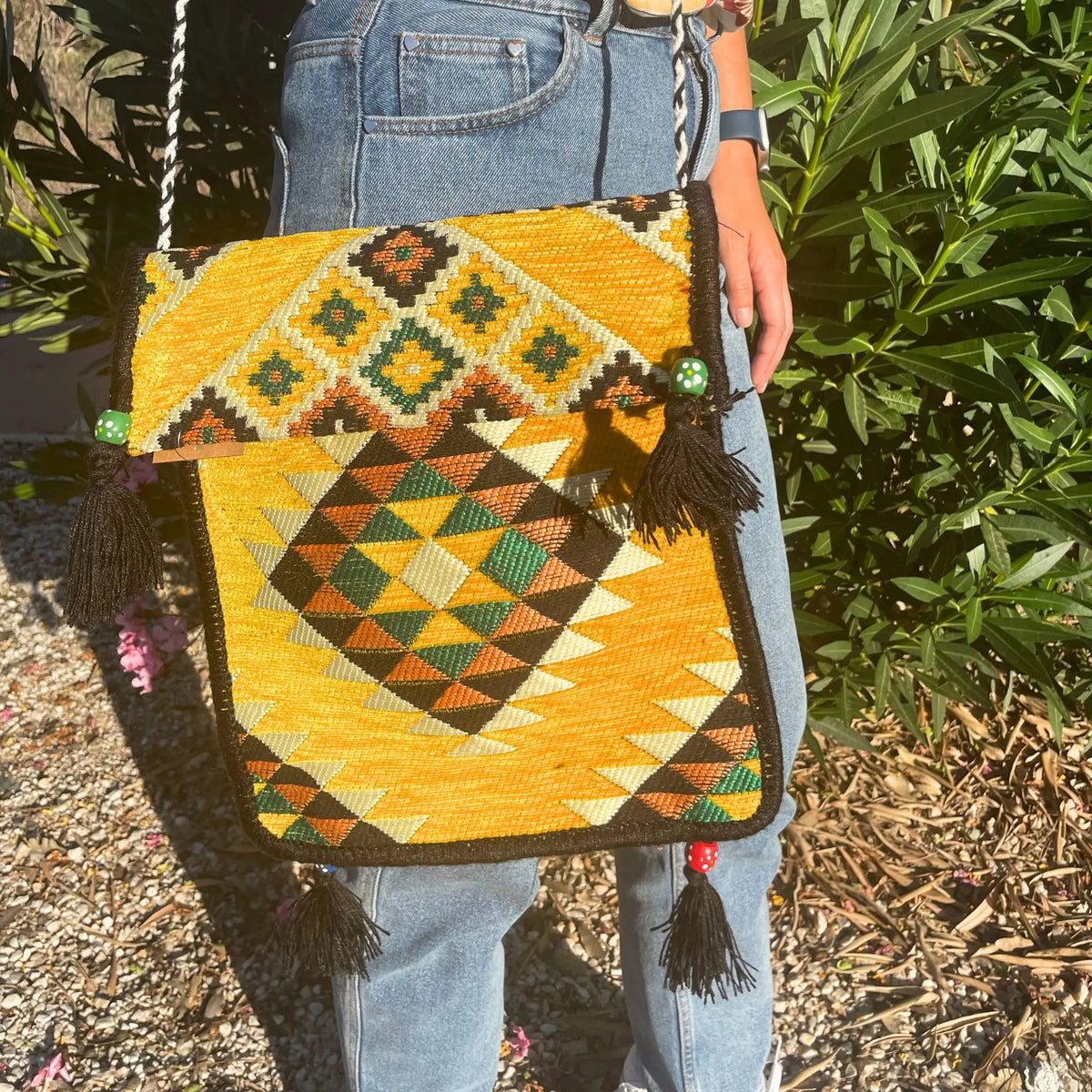 Bolsa mensajera de tela tipo kilim, diseño geométrico amarillo, verde y negro con borlas y cuentas decorativas.