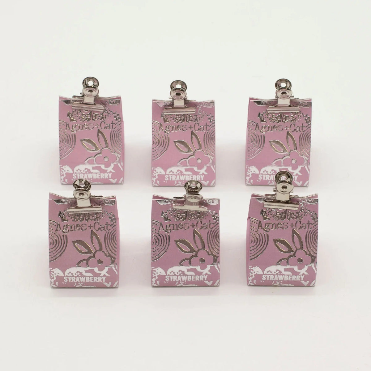 Seis bálsamos labiales Agnes + Cat de fresa en bolsas de papel rosa con pinzas metálicas sobre fondo blanco.