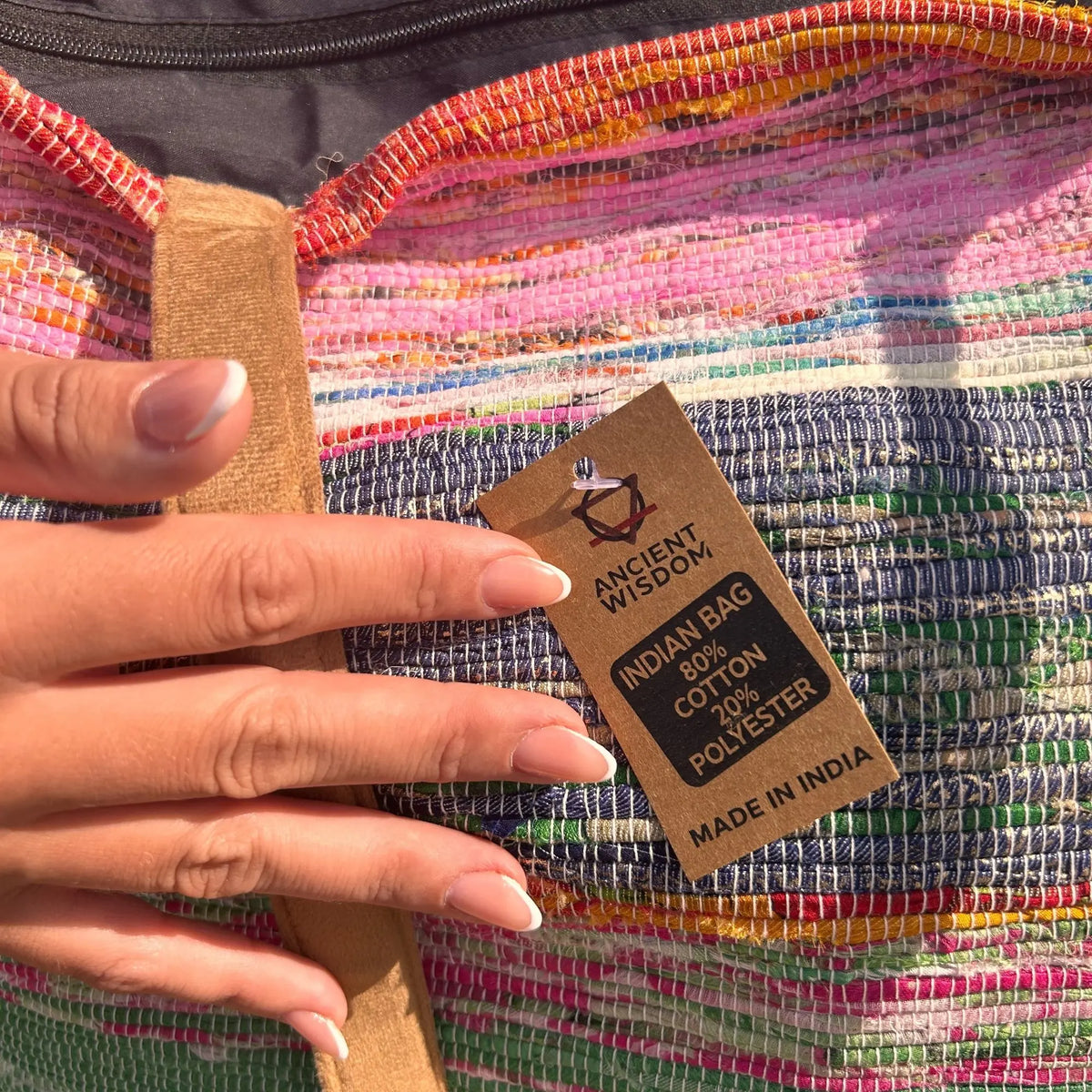 Bolso de tejido artesanal en tonos rosas y azules con correa de tela, mostrando una etiqueta de composición textil.