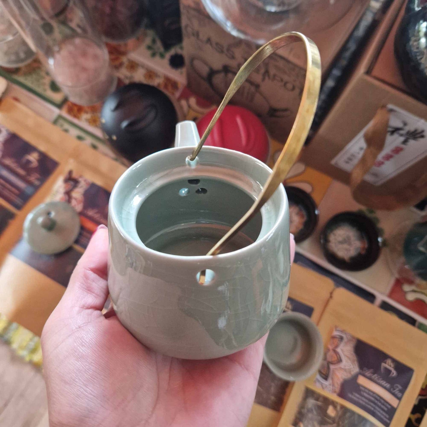 Tetera de cerámica verde con acabado craquelado y asa metálica dorada, sostenida en una mano. Marca Ancient Wisdom.