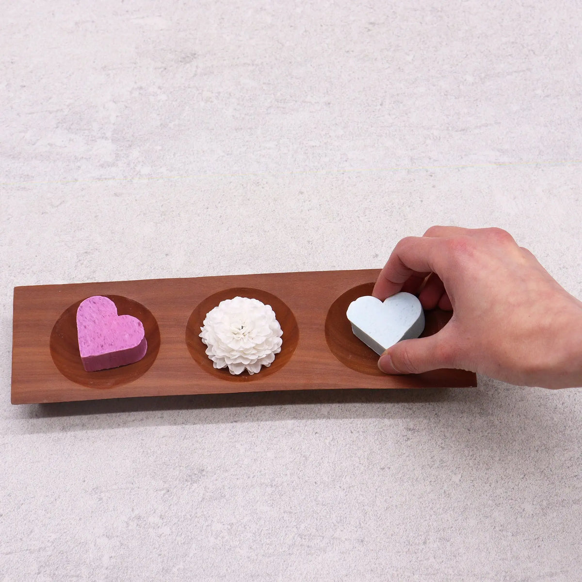 Jabonera rectangular de madera con tres compartimentos circulares; contiene dos jabones en corazón y una flor blanca.