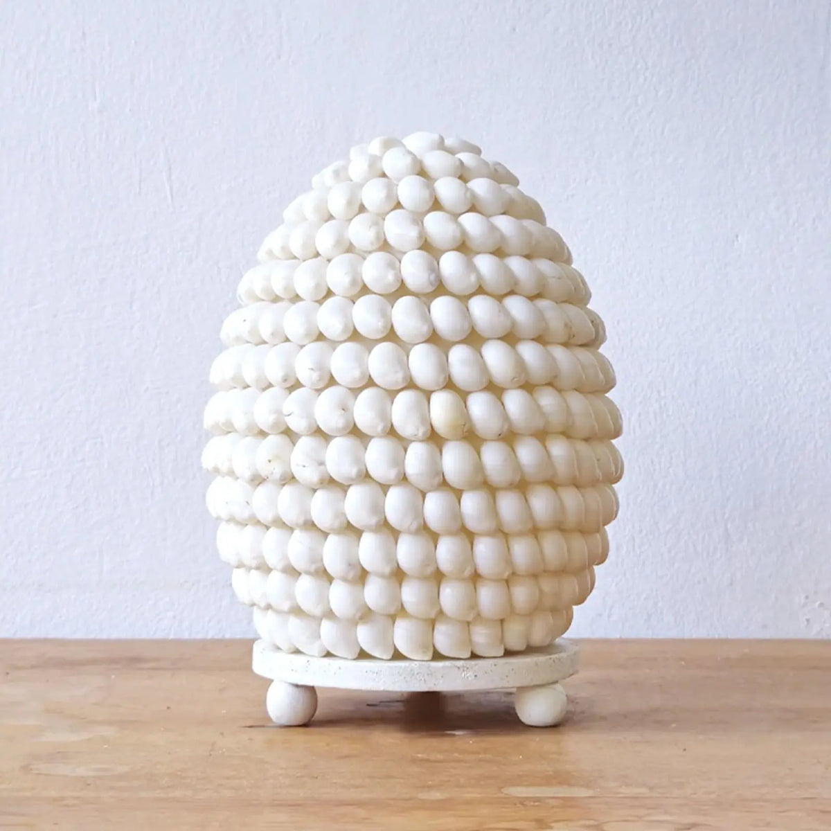 Lámpara ovalada estilo boho de conchas blancas naturales sobre una pequeña base con patas, fondo blanco.