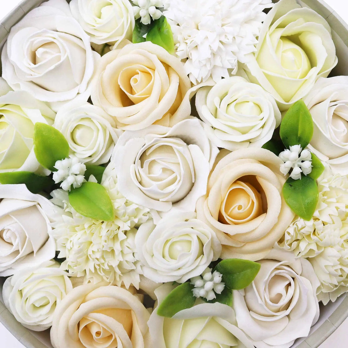 Caja redonda con flores de jabón en tonos blanco y marfil, decoradas con hojas verdes y pequeñas flores blancas.