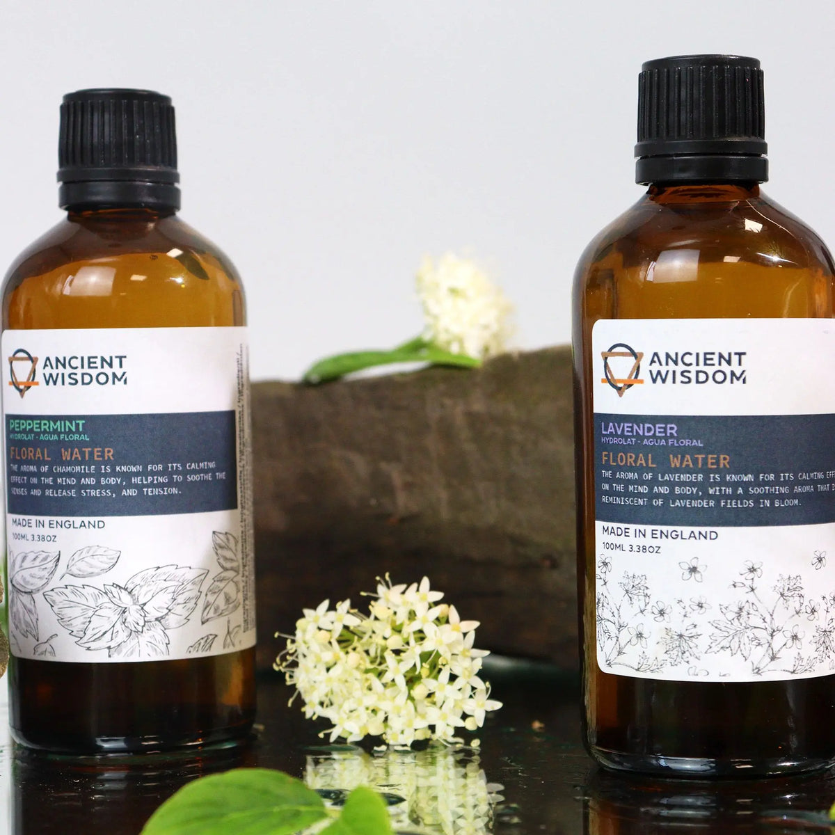 Dos botellas de vidrio ámbar con etiquetas blancas e ilustraciones botánicas, sobre una base oscura con flores blancas.