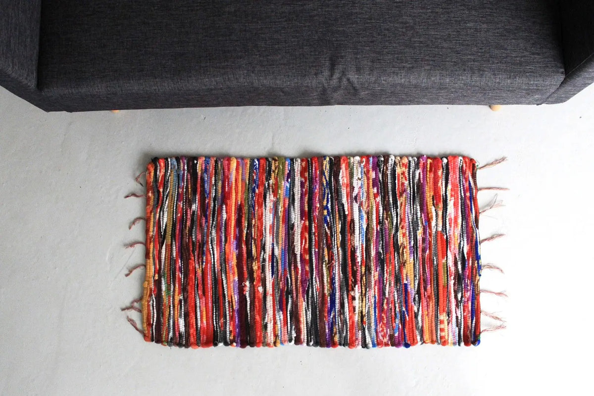 Alfombra rectangular artesanal de trapos multicolor, tejido de tiras de tela texturizadas con flecos en los extremos.