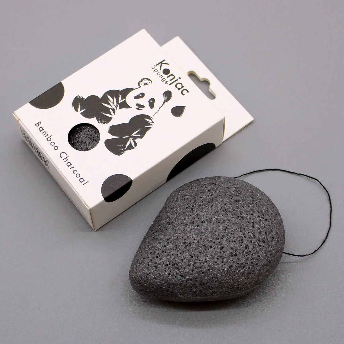 Esponja facial de carbón, porosa y negra, con forma de gota y cordón, junto a su caja blanca ilustrada con un panda.