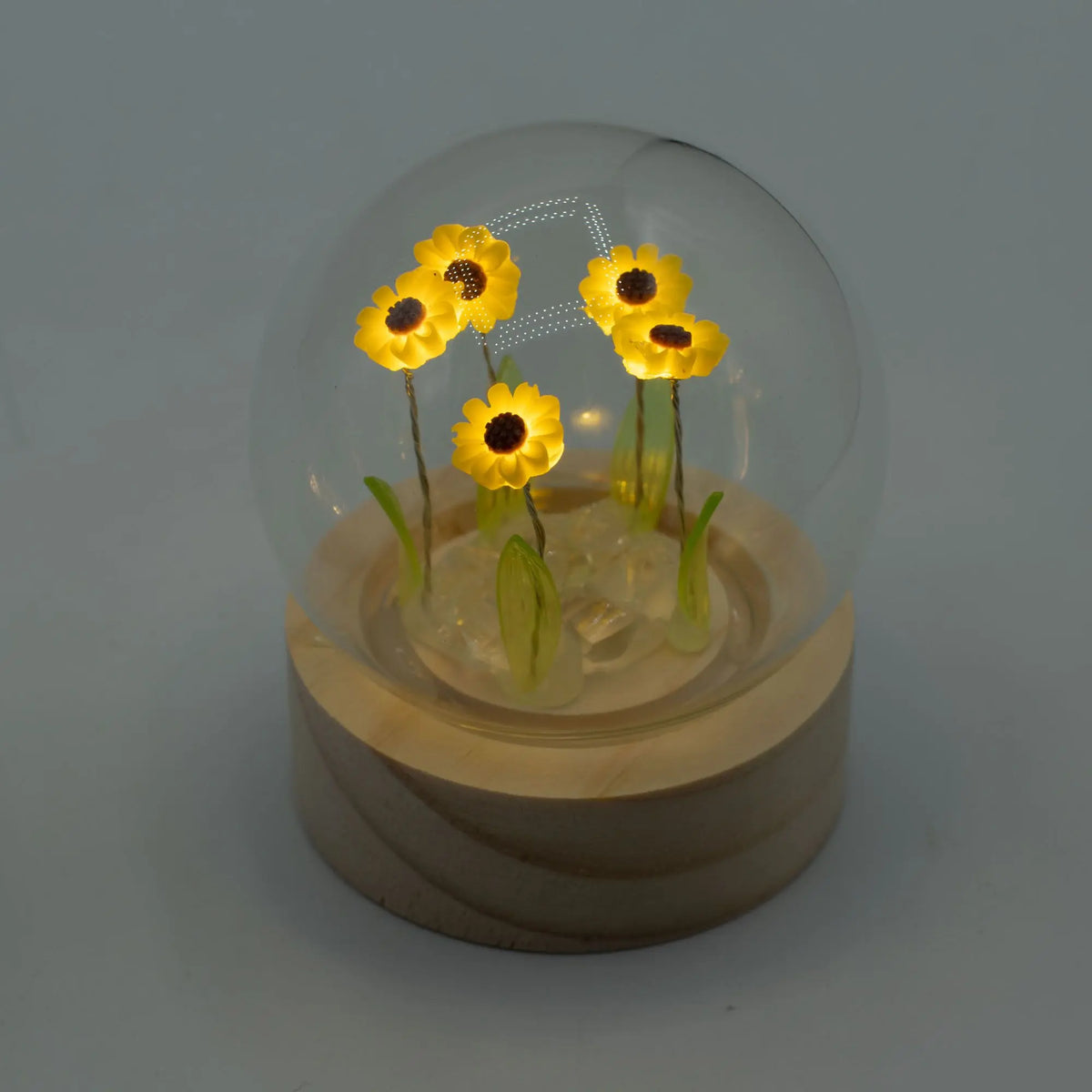 Domo de cristal con 5 girasoles LED iluminados, sobre base circular de madera clara. Diseño floral brillante y elegante.