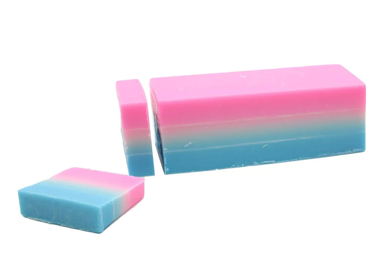 Jabón artesanal rectangular degradado en rosa y azul, con una rodaja cortada, sobre fondo blanco liso.